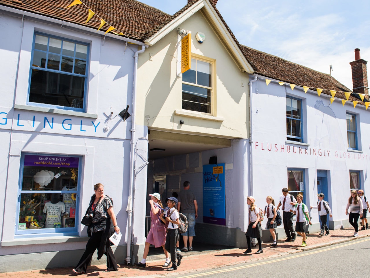 A line of exciting school children in school uniform walk past the Roald Dahl Museum. It's a bright, sunny day and they are being led by a teacher. The Museum building behind them is painted light yellow and purple and has jolly yellow bunting on the roof.