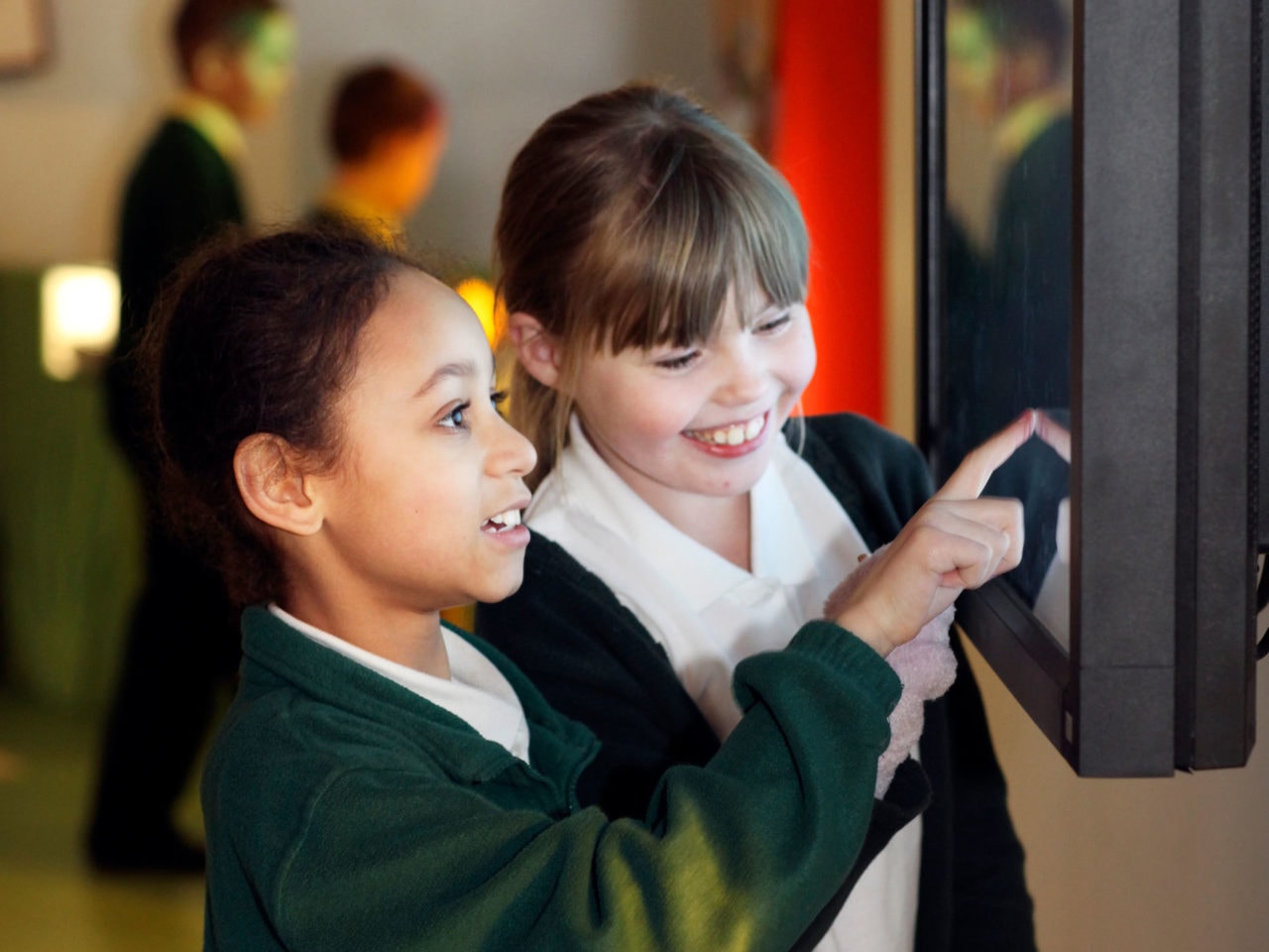 Two primary school children look up at a touch screen mounted on a wall. They are both looking and the screen and smiling. One girl has her finger on the screen.