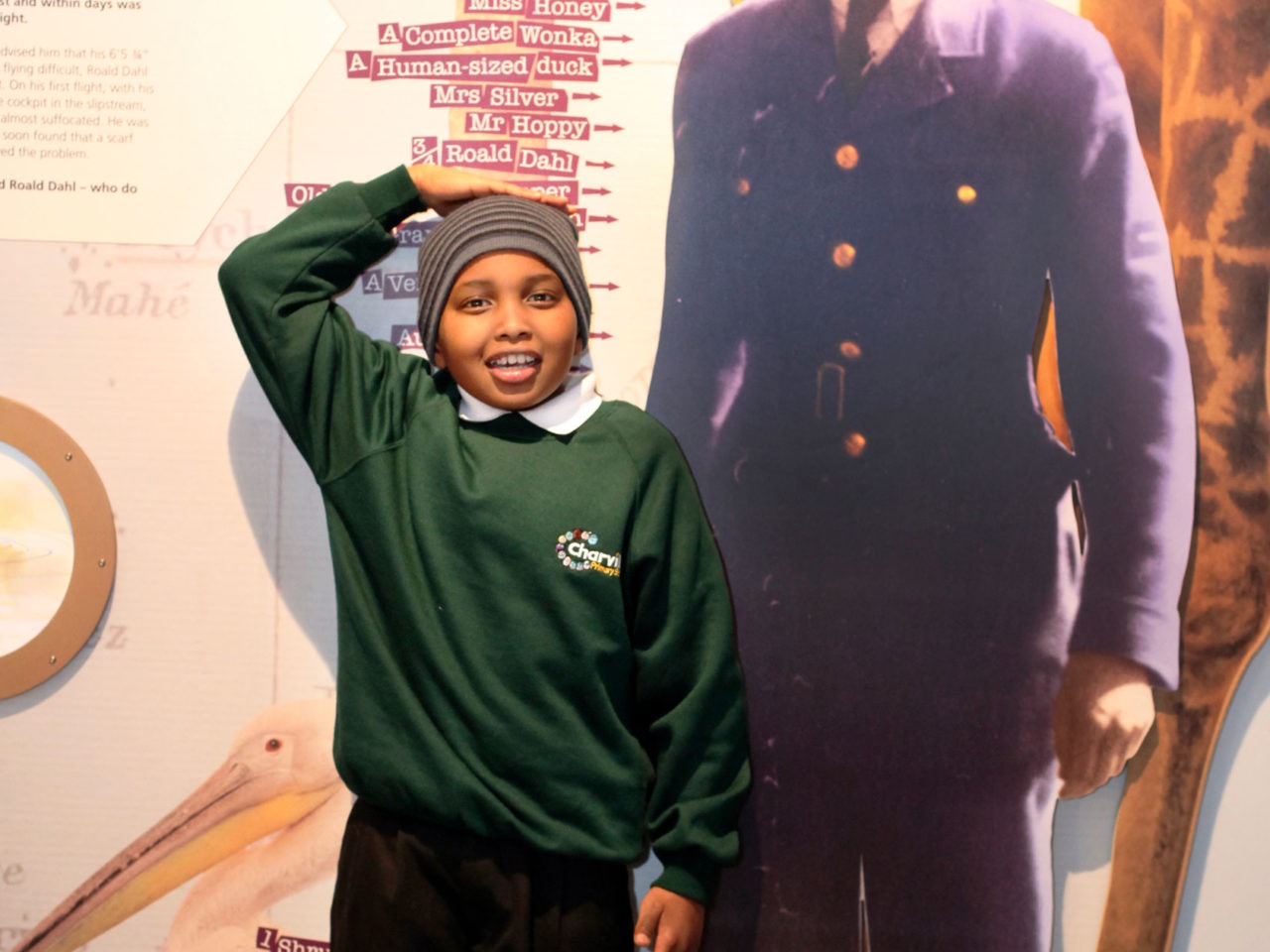 A photo of a smiling boy. He's in green school uniform and stood against a height chart that features the heights of Roald Dahl characters. He is holing his right hand on top of his head to measure himself.