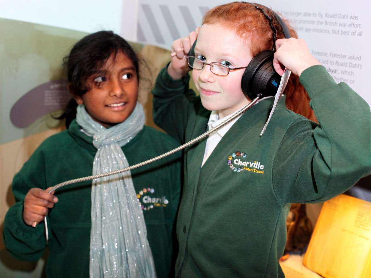 Two children in green school uniform in a museum gallery. One has headphones on her head and is holding them The other child is looking up towards her friend.