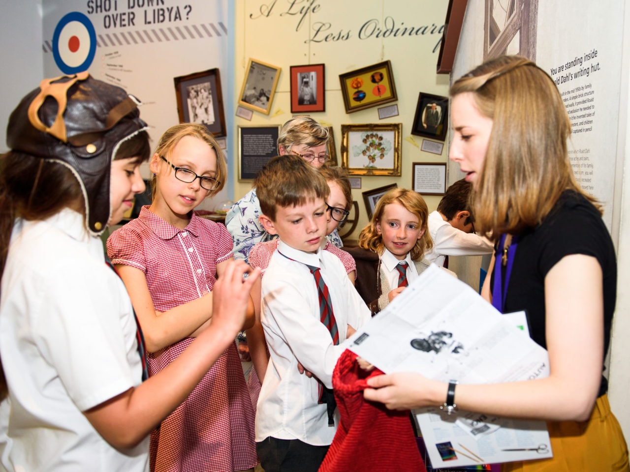 A group of excited primary school children gather round an adult school session leader. They are in a museum gallery with lots of colourful graphics on the wall. The session leader is handing something out to the children.