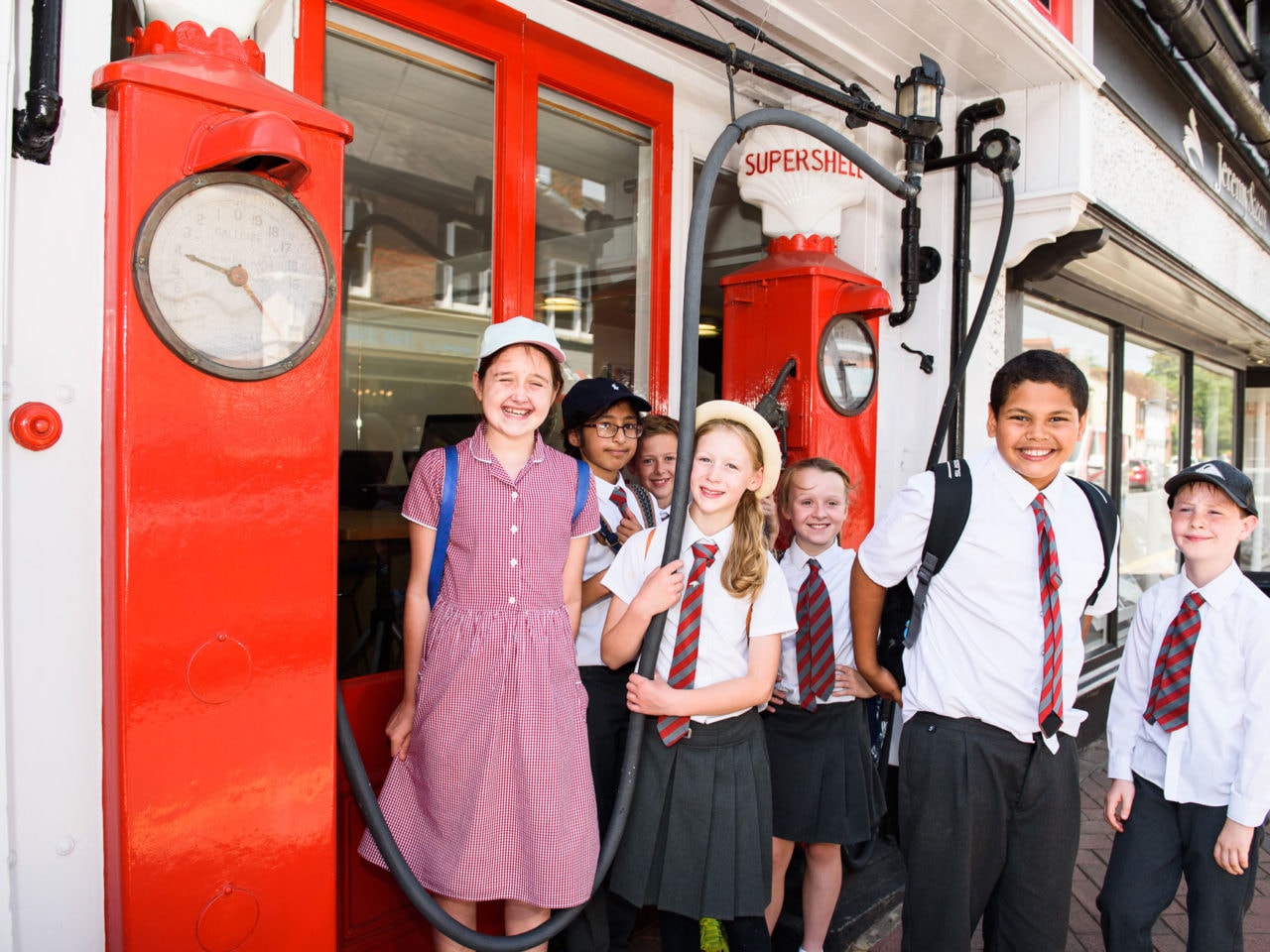 Seven primary school children are standing in a group around an old petrol pump. The bright red pumps have 'Supershell' written on the top. The kids are in school uniform and looking at the camera smiling. It's a nice, sunny day.