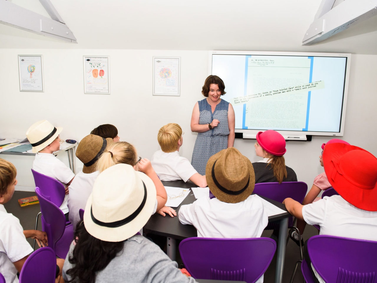 A school session leader faces the camera. In front of her are a group of primary school children sat in chairs. They have their backs to the cameras and they all have different, colourful hats on. They are engrossed in what the adult is saying.