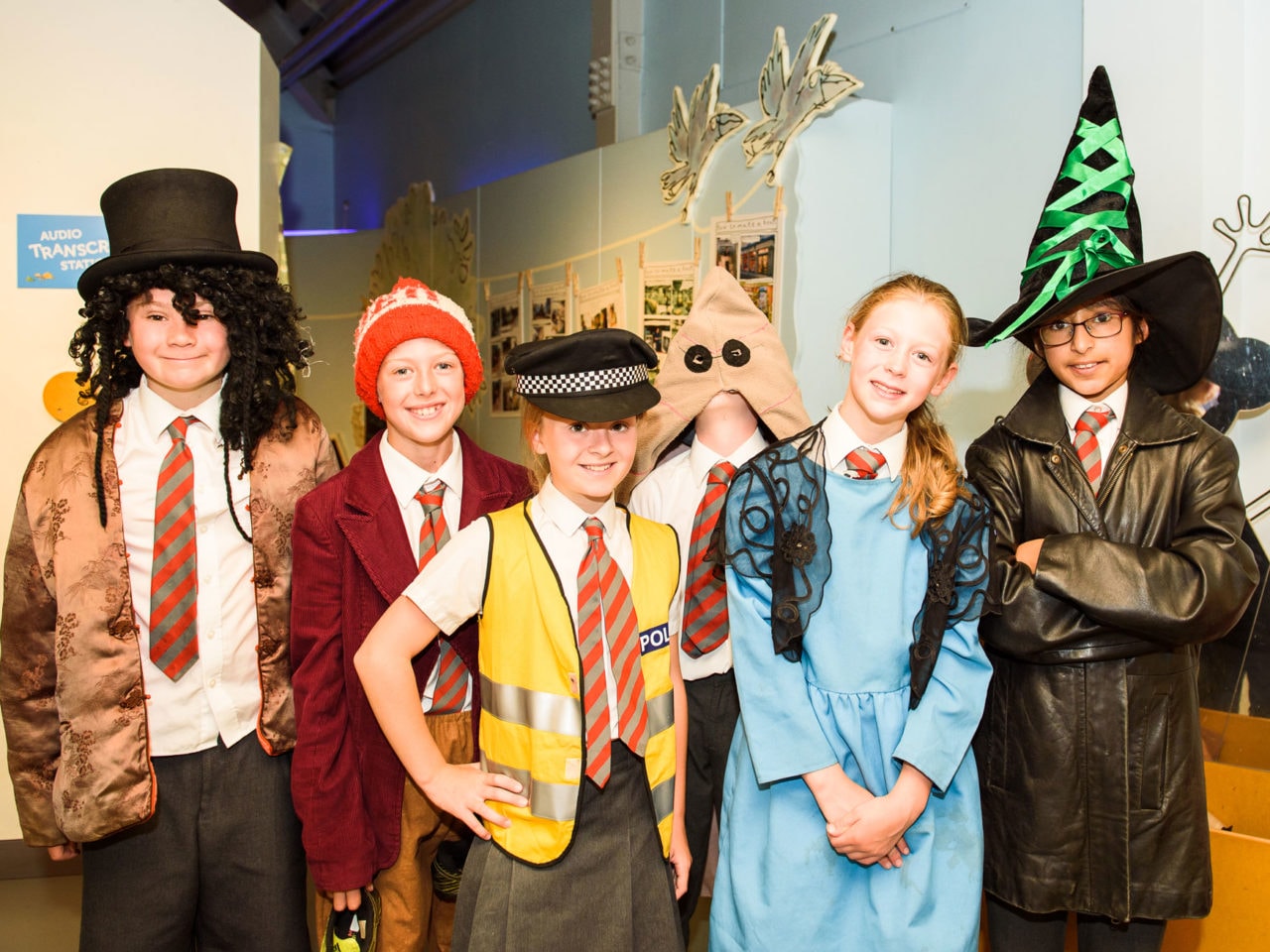 Six children stand facing the camera. They have all been trying on some fancy dress costumes and have various garments over their school uniform. One has a witches hat, one has a blue dress, one looks like a police woman, other are wearing interesting hats.
