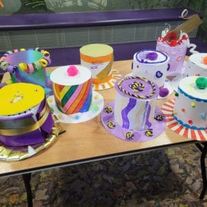 A selection of handmade top hats. They are bright an colourful and covered in ribbon, pipe cleaners and pop-poms. The are all sat on a wooden table