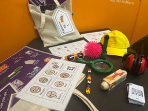 Some of the contents of our SEND explorers kits on a dark grey table. There are red ear defenders, a yellow cap, a green magnifying glass, fidget toys and emotion cards