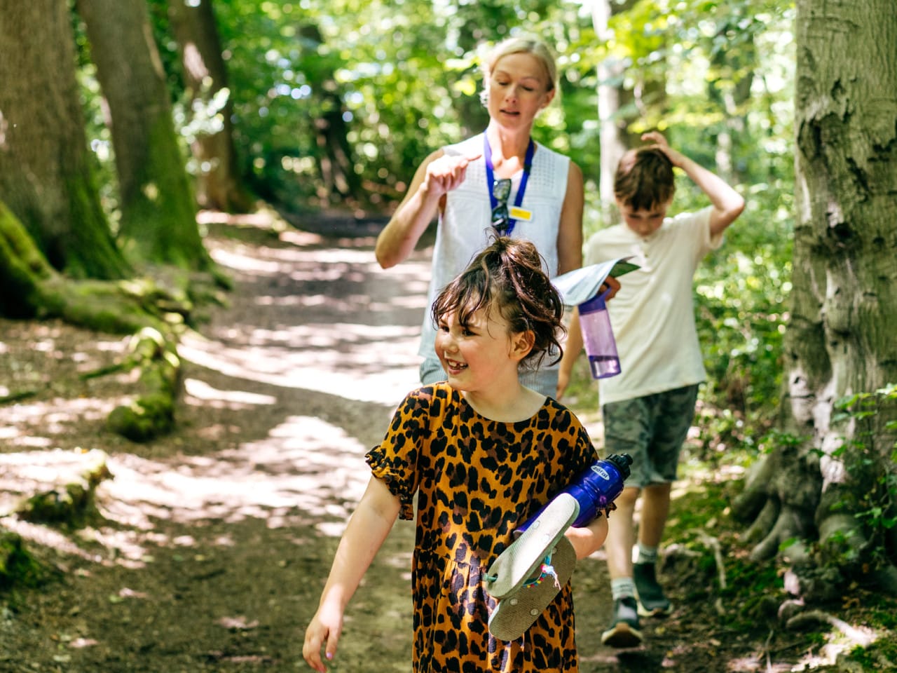 A group of people walking in the woods in summer. They are in dappled shade and the sun brraking through makes it look like it miust be a very sunny day. A small girls walks in the foreground smiling, with a woman and boy talking together as they walk behind her.